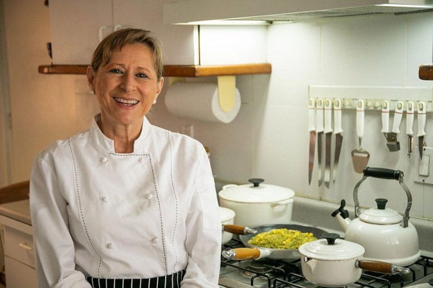 A chef in a white uniform standing in a kitchen with a stove, pots, and a hanging knife rack. 3