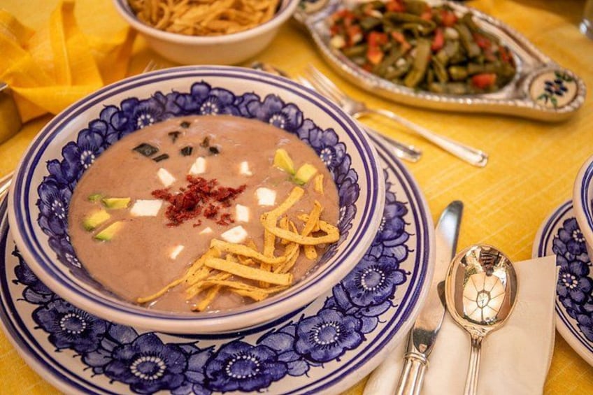 Un tazón de sopa con un borde azul decorativo, acompañado de una cuchara y una servilleta sobre un mantel amarillo. 2