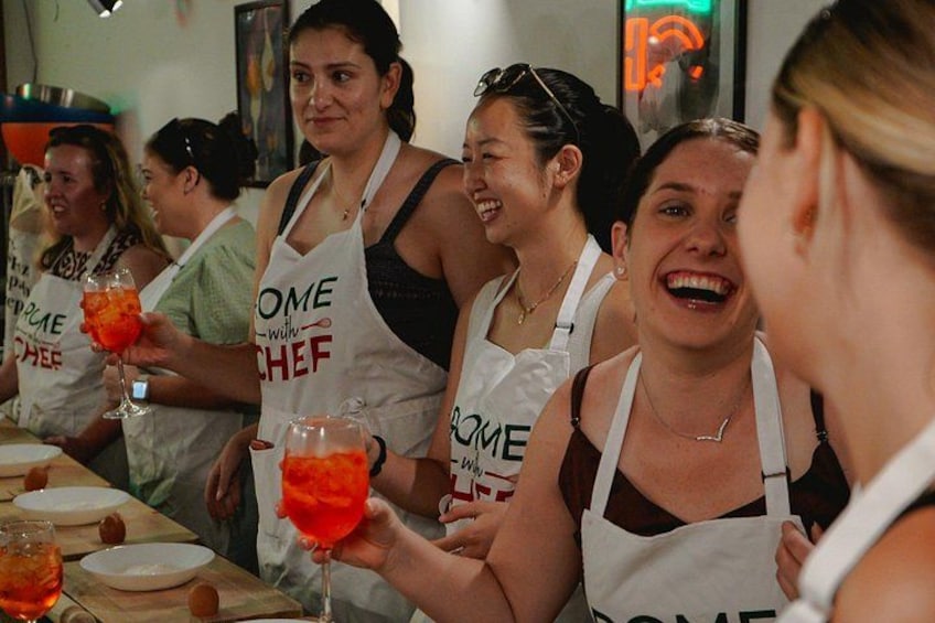 Un gruppo di persone con grembiuli con la scritta 'HOME CHEF', che tengono bicchieri con liquido rosso, in una cucina. 2