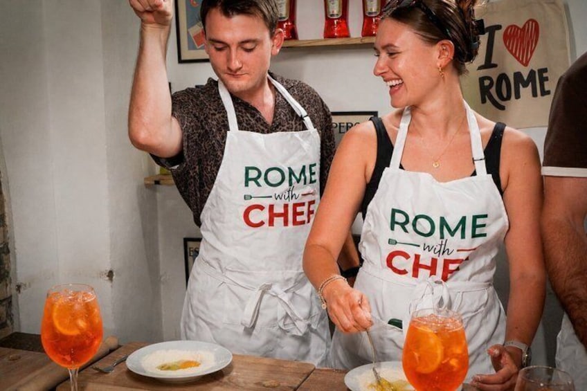 Zwei Personen in Schürzen mit dem Aufdruck 'ROME with CHEF', ein Tisch mit Tellern und Gläsern sowie ein Schild im Hintergrund mit der Aufschrift 'I ♥ ROME'. 1