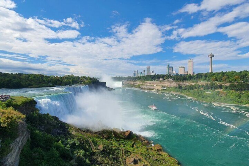 A waterfall, river, and city skyline with greenery and a boat. 1