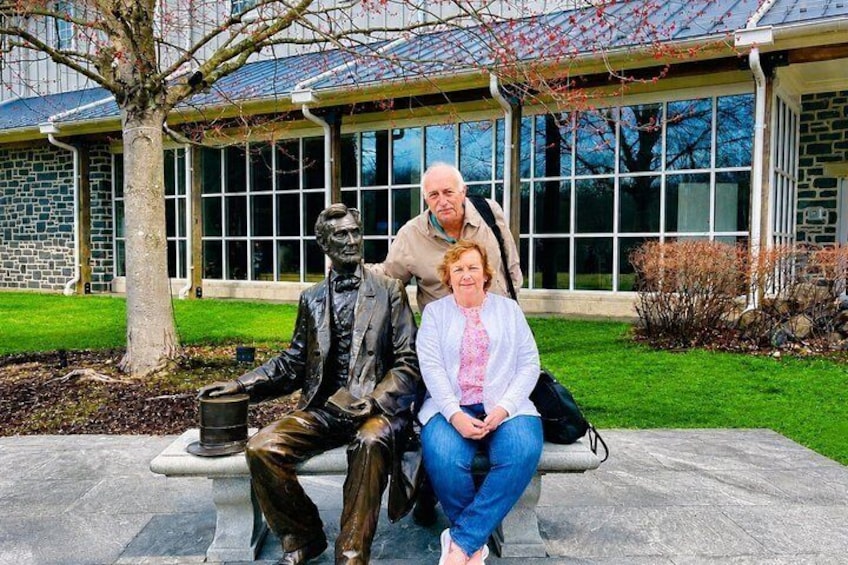 A statue of a seated man and woman on a bench in front of a building with large windows and a stone facade. 1