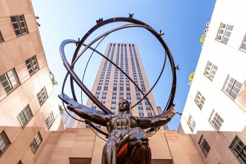 A sculpture of a figure with outstretched arms and a circular element behind it, set against a backdrop of tall buildings. 2
