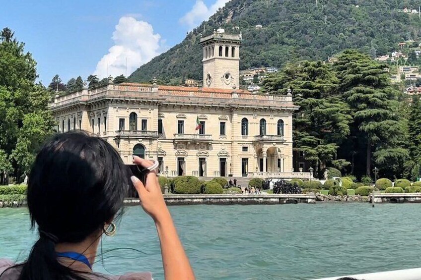 A person taking a photo of a historic building with a tower, surrounded by water and greenery. 1