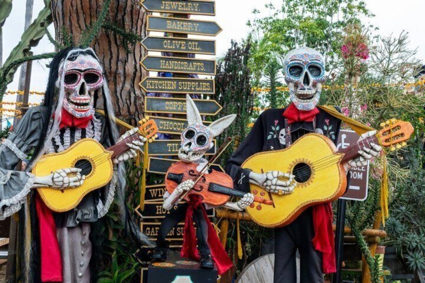 Skeleton figures dressed in traditional attire, holding guitars, with a signpost listing items like jewelry and bakery. 1