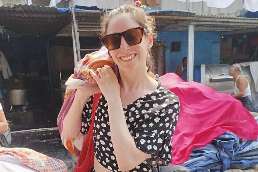 A person holding a pink cloth, standing in an outdoor market with visible stalls and a blue building in the background. 1