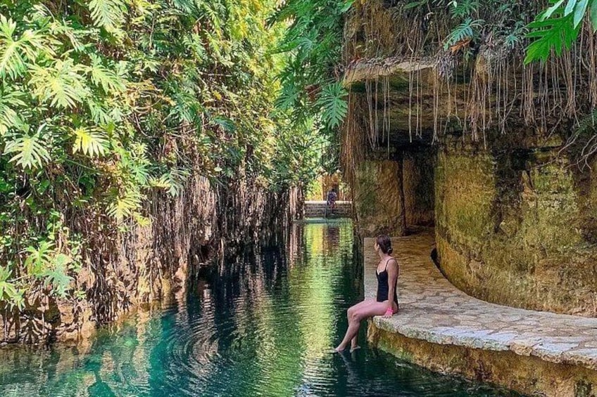 A person sitting by a calm water body surrounded by lush greenery and a stone structure. 1