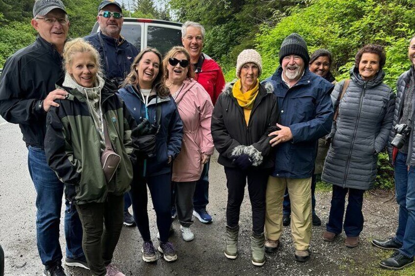A group of people standing outdoors, some wearing winter clothing, with a vehicle and greenery in the background. 5