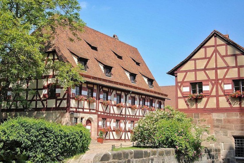 A traditional half-timbered house with a steeply pitched roof, surrounded by greenery and a stone wall. 1