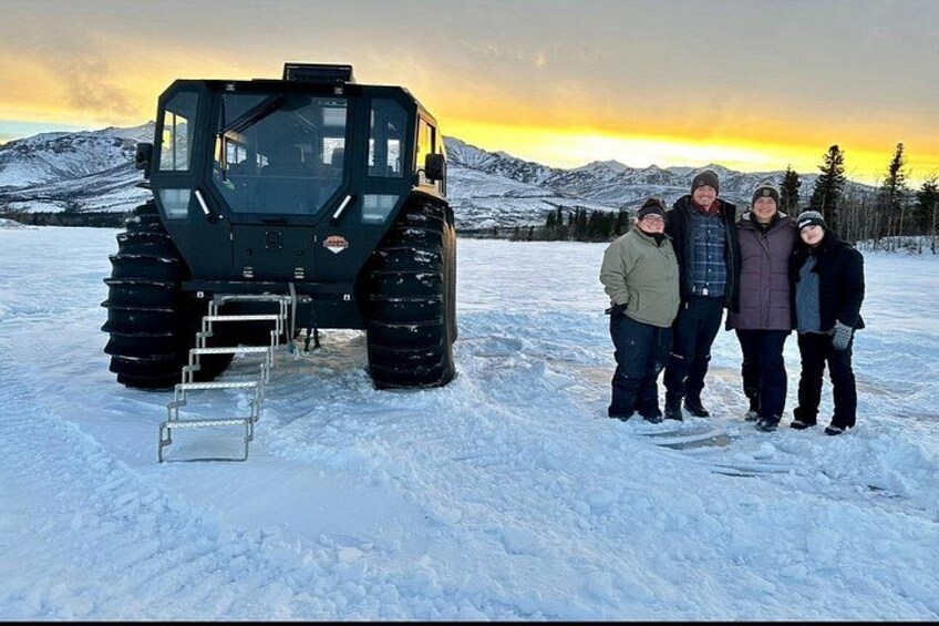 Un groupe de personnes se tient dans un paysage enneigé, avec un grand véhicule en arrière-plan. 2