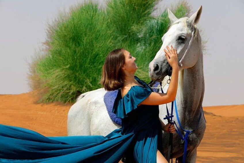 Una persona con vestido azul junto a un caballo en un desierto con dunas de arena y vegetación verde. 4