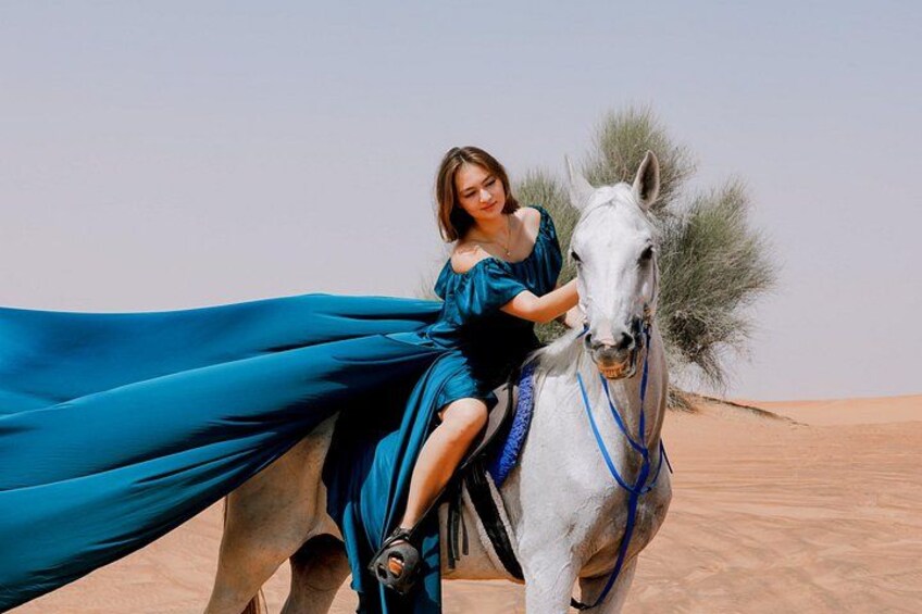 Una persona vestida de azul montando un caballo blanco en un desierto con dunas de arena y vegetación escasa. 3