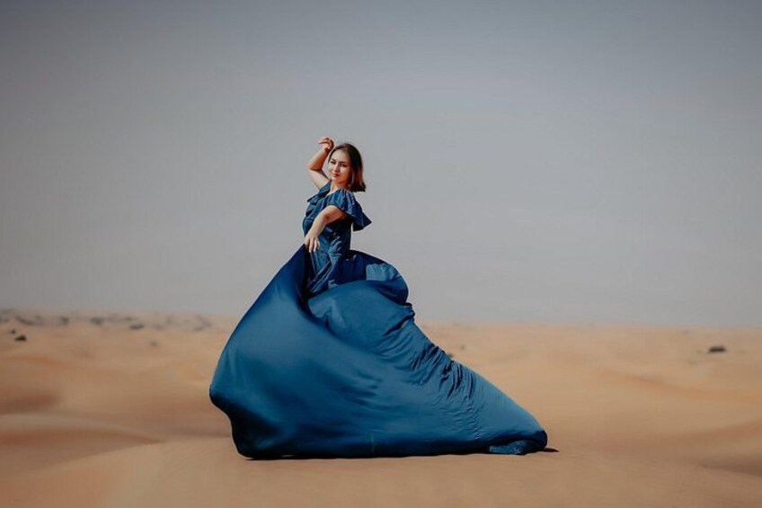 Una persona sentada sobre una duna de arena en un paisaje desértico, vestida con un vestido azul y suelto. 5