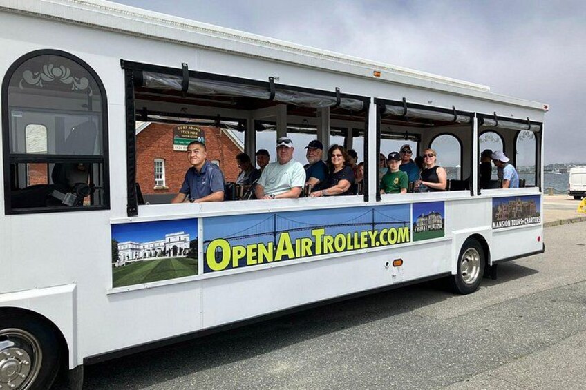 A white trolley with passengers, a sign reading 'OPENAIRTROLLEY.COM', and a brick building in the background. 1