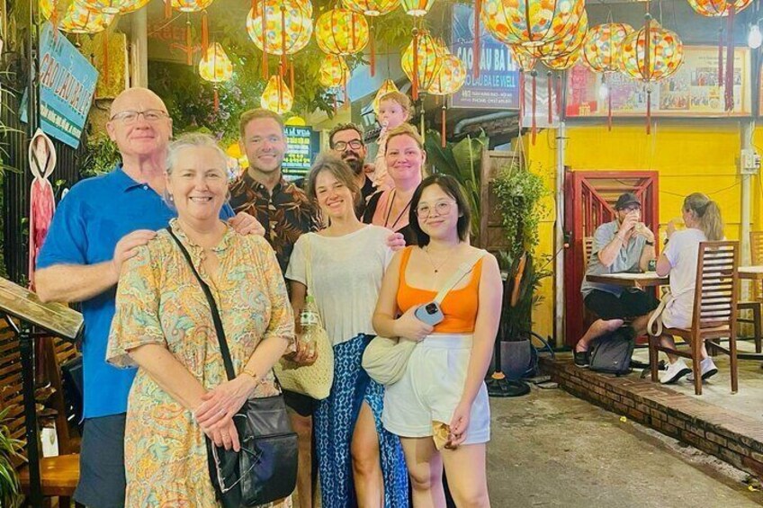 A group of people posing in a brightly lit outdoor dining area with hanging lanterns and a yellow wall. 2