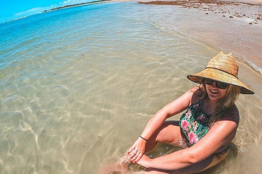 A person sitting on a wooden float in clear shallow water, with a beach and ocean in the background. 5