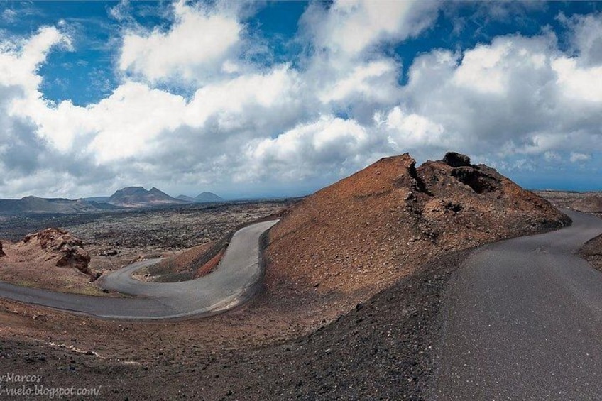 Une route sinueuse traverse un paysage aride, parsemé de rochers, avec des montagnes lointaines qui se dessinent sous un ciel partiellement nuageux. 2