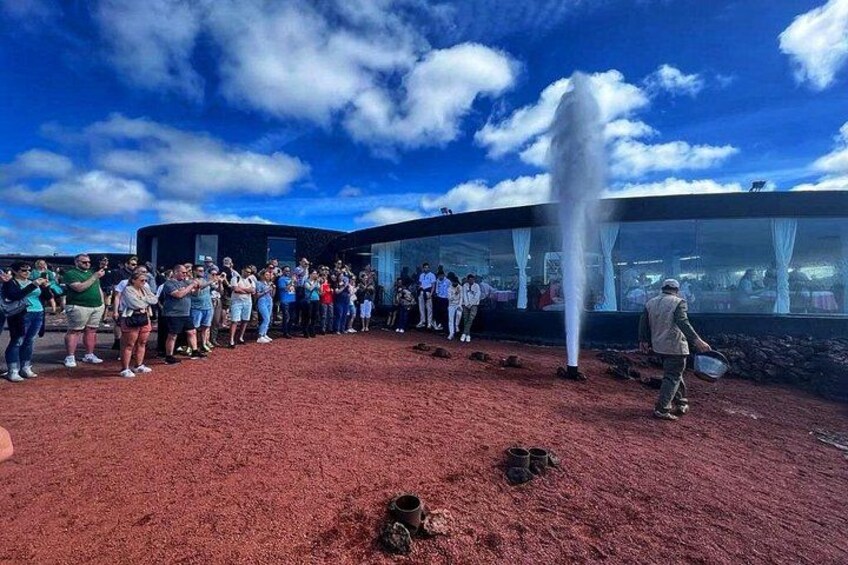 Un groupe de personnes se tient sur une étendue de sable rouge, près d’une fontaine et d’un bâtiment moderne aux murs de verre. 3