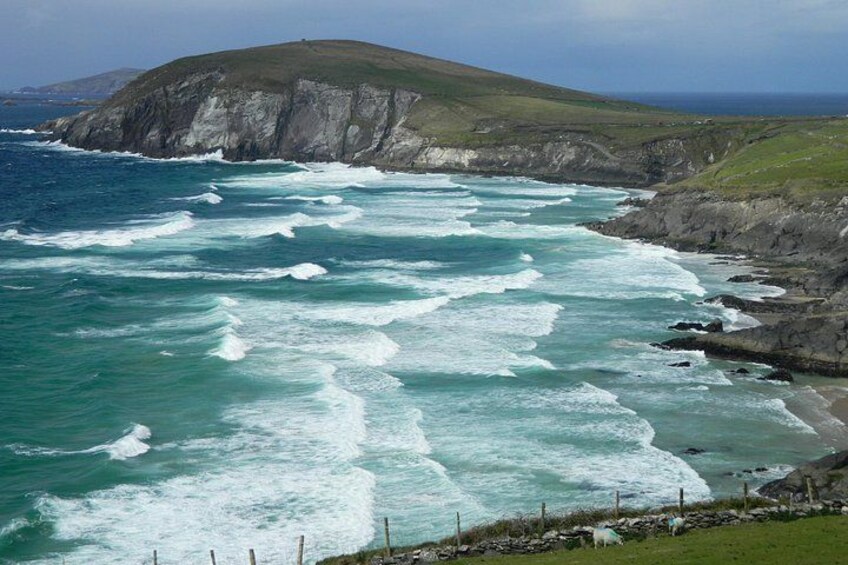 Paisaje costero con olas rompiendo contra una costa rocosa, una colina cubierta de hierba al fondo y una valla a lo largo de la playa. 2