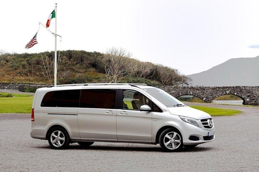 A white minivan parked on a gravel road with a stone bridge and flags in the background. 2