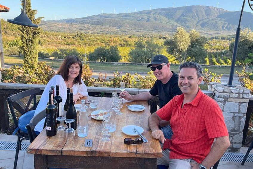 Three people seated at an outdoor table with wine bottles and glasses, set against a scenic vineyard backdrop. 3