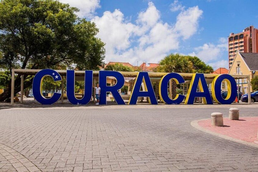 Store blå bogstaver der staver CURACAO på en belagt overflade med træer og bygninger i baggrunden. 1