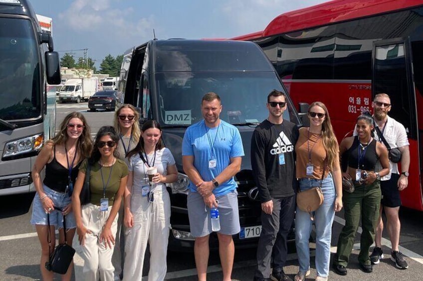 A group of people standing in front of a black van with the license plate DMZ. 5