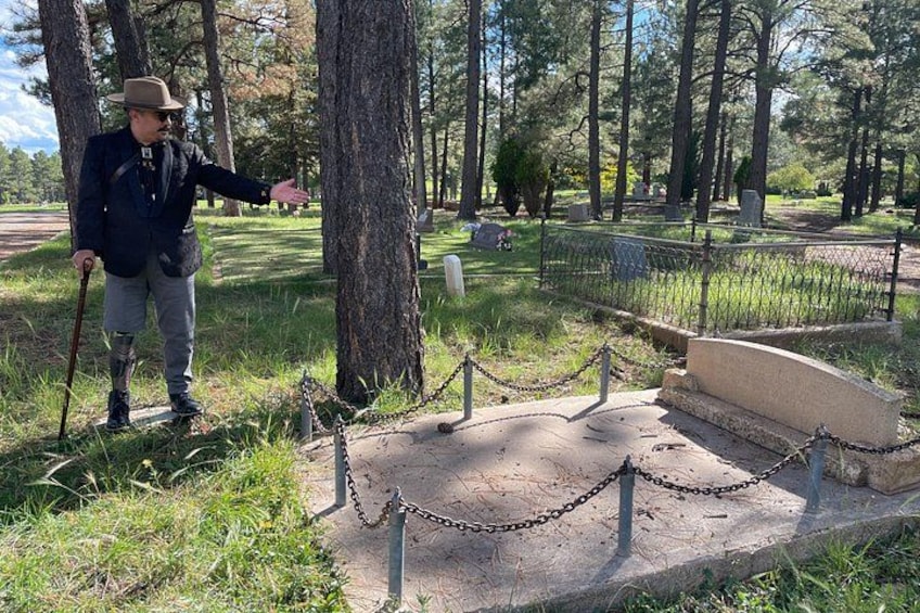 Un hombre con vestimenta de época está de pie junto a un cementerio con una cerca de malla y lápidas. 3