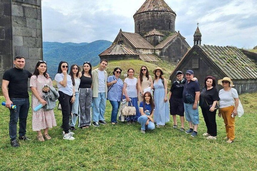 A group of people posing in front of a historical building with a dome and a bell tower, surrounded by a mountainous landscape. 1