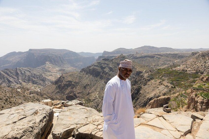 A man in traditional attire standing on rocky terrain with a mountainous landscape in the background. 5