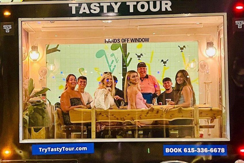 Un ristorante Tasty Tour con un cartello che recita 'NON TOCCARE LA FINESTRA' e un gruppo di persone all'interno. 3