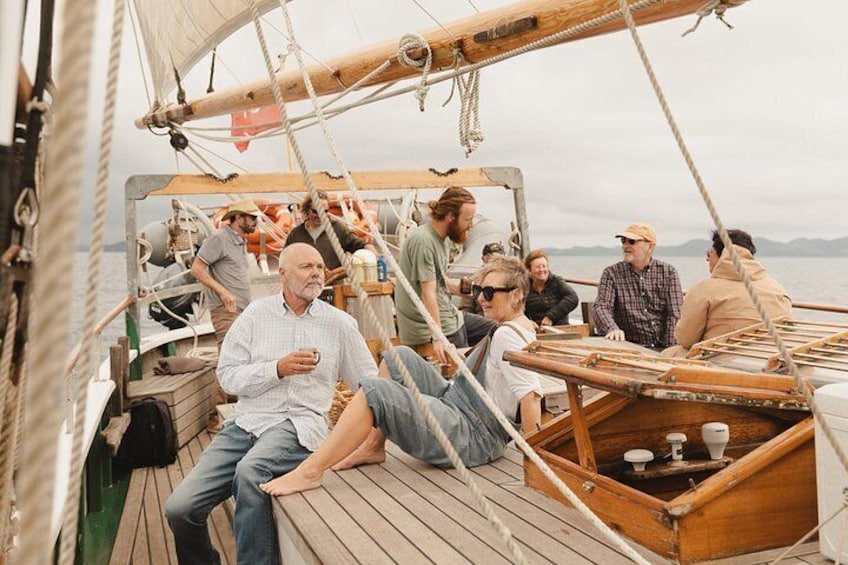 A wooden deck on a boat with people sitting and standing, a small wooden structure with a toilet, and a rope netting. 3