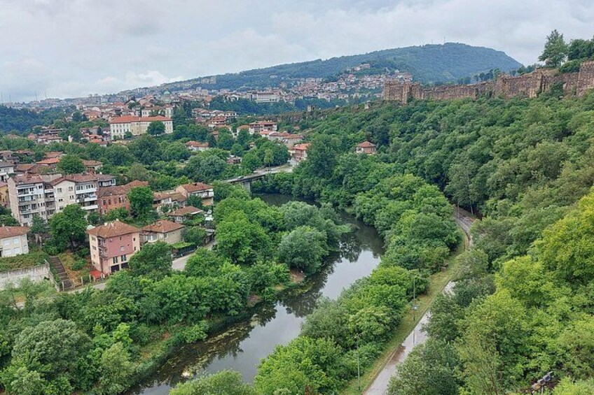 Un río serpentea a través de una zona boscosa densa, con edificios a la izquierda y una colina con ruinas al fondo. 2
