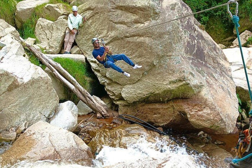 Two people sitting on rocks by a flowing stream, with a rope bridge in the background. 3