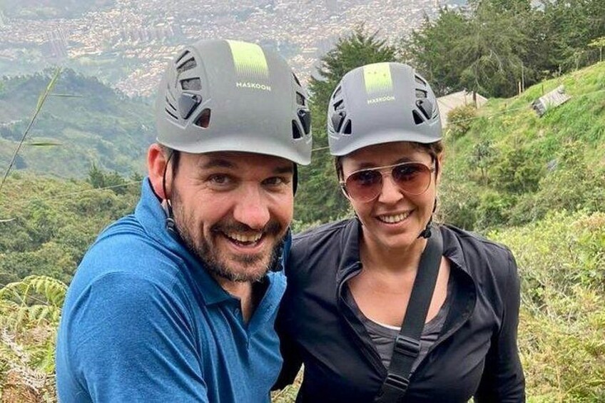 Two people wearing helmets with 'HASKON' on them, standing on a grassy hill with a view of a town in the background. 2