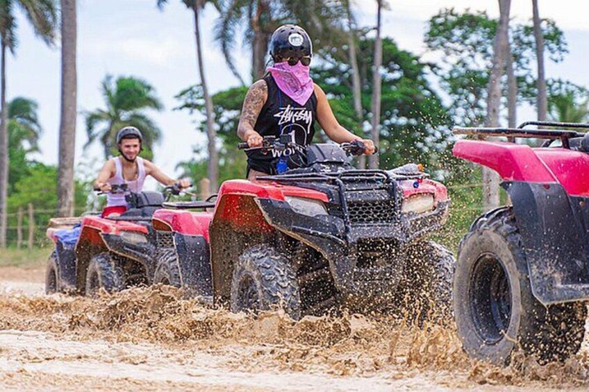 Three people riding ATVs on a muddy path with palm trees in the background. 1