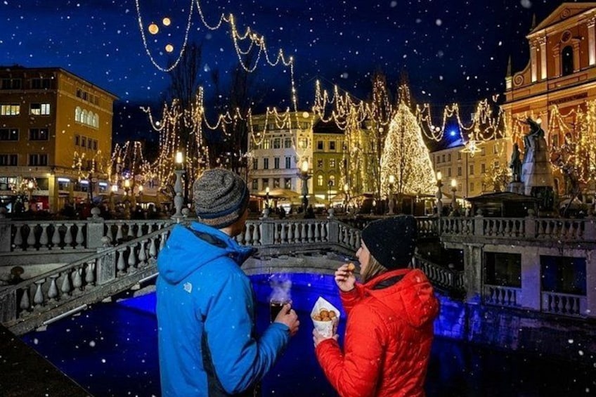 Two people standing by a bridge with a festive background of illuminated trees and buildings. 3