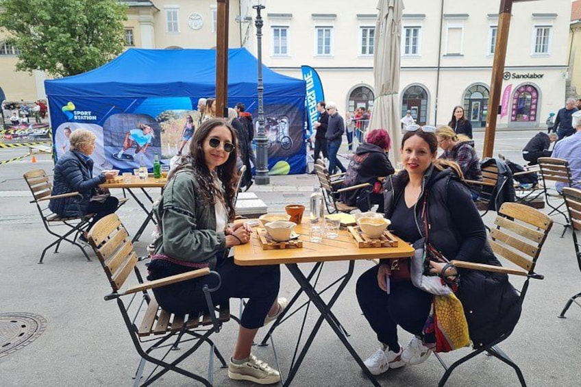 Two people sitting at a wooden table outdoors with a blue tent and other people in the background. 2