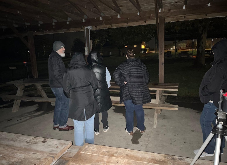 Un gruppo di persone in piedi sotto una tettoia di notte, con un tavolo da picnic e delle panchine in vista. 1