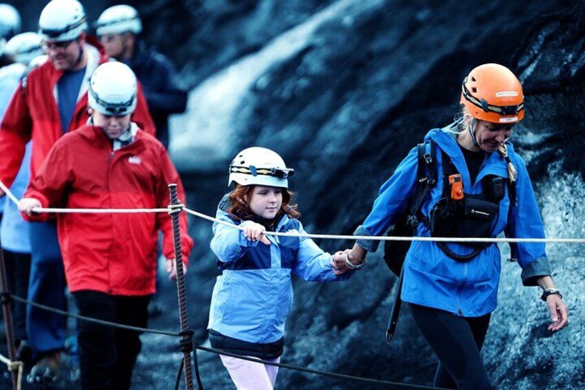 A group of people in outdoor gear, including helmets and backpacks, walking on a rope bridge over a rocky terrain. 1