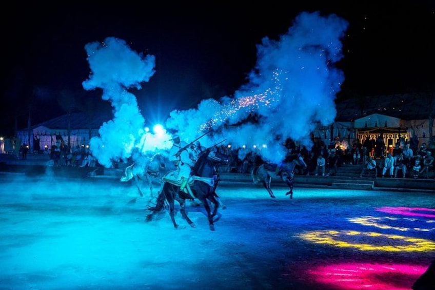 A nighttime outdoor event with horses and riders, illuminated by blue and pink lights, and smoke effects. 1