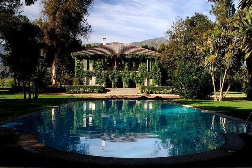 A swimming pool surrounded by a lush garden, a house with a tiled roof, and a mountainous backdrop. 1