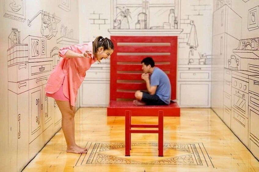 A person in pink bending over a red bench, another person sitting on it, and a kitchen-themed wall in the background. 3