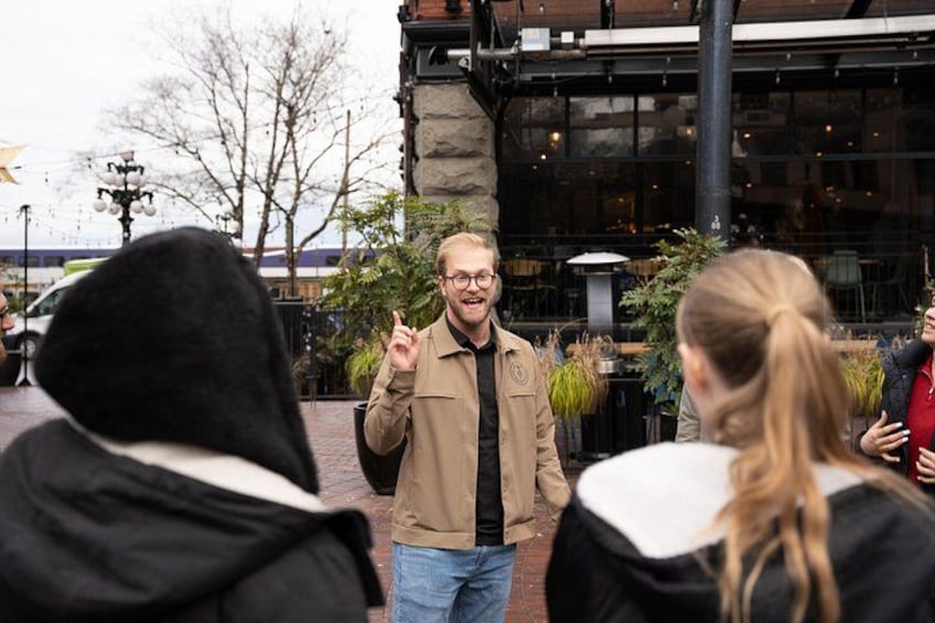 A person in a brown jacket gesturing while speaking to a group of people outdoors. 5