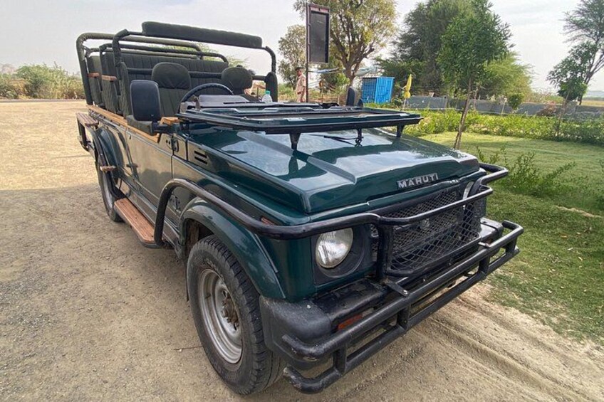 A green Maruti vehicle parked on a dirt path with a grassy field and trees in the background. 2