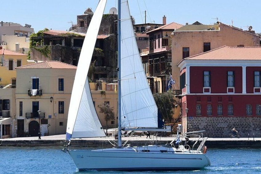 A sailboat with white sails on calm water, surrounded by colorful buildings. 1