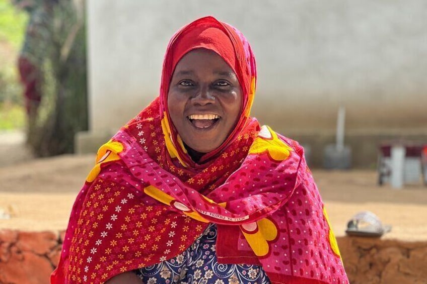 A person wearing a red and yellow patterned shawl, seated outdoors with a concrete structure and a tree in the background. 1