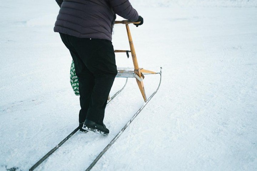A person standing on a sled with a wooden frame, wearing black pants and a purple jacket, on a snowy surface. 3