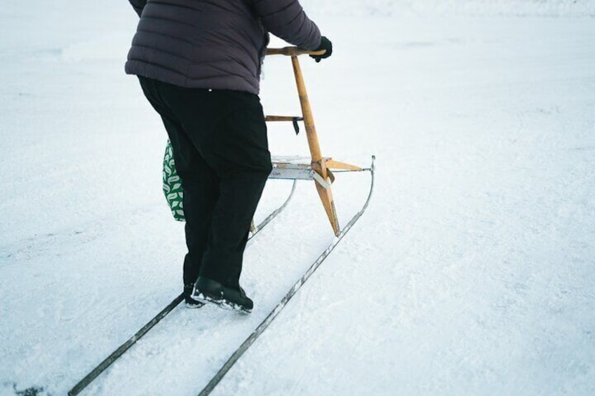 A person standing on a sled with a wooden frame, wearing black pants and a purple jacket, on a snowy surface. 5