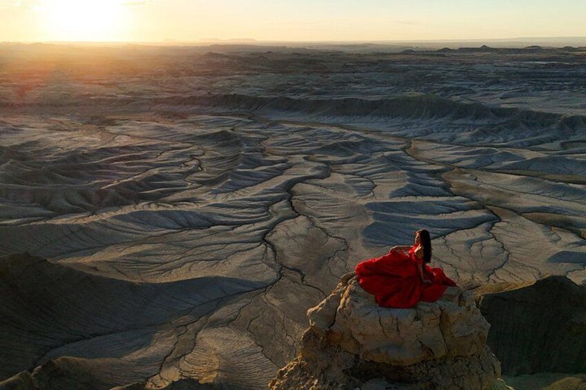 Une personne vêtue d’une robe rouge est assise sur une formation rocheuse, dans un vaste paysage craquelé, au coucher du soleil. 3